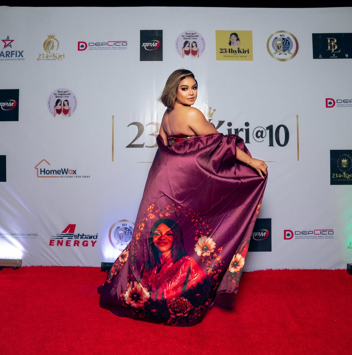 Tessy Kiri Bibowei on the 234byKiri@10 red carpet in a magenta floral cape printed with a family portrait